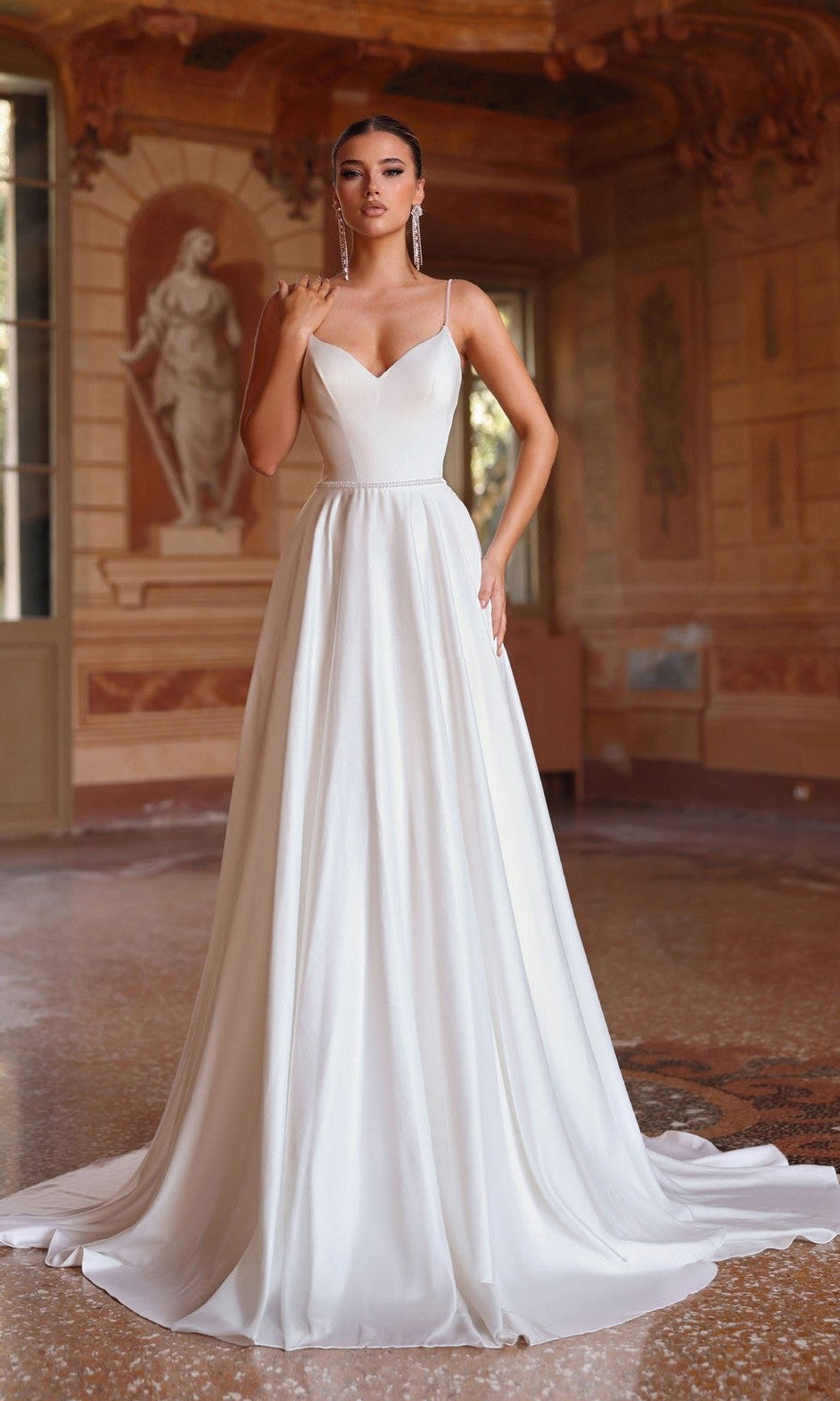 A woman in the Alyce 7099 Long White Dress, featuring beaded spaghetti straps and a fitted bodice in diamond white, stands in an ornate room with statues and detailed wall art.